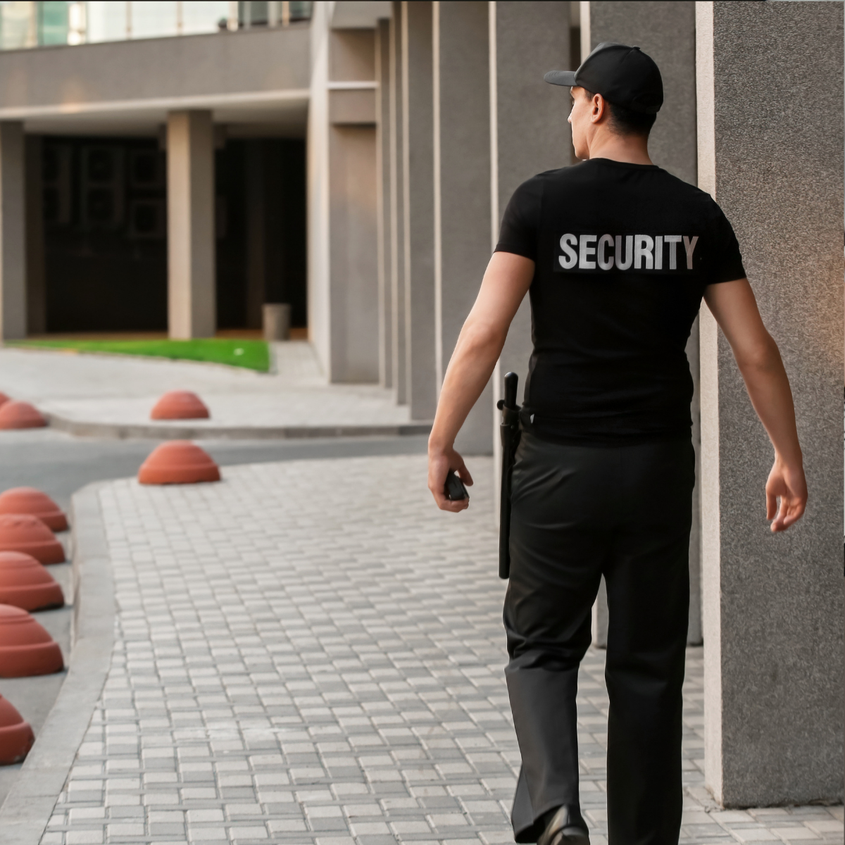 Un agent de sécurité en uniforme sombre, avec l’inscription « SECURITY » dans le dos, surveille l’entrée d’un immeuble moderne. Un agent de sécurité en uniforme sombre, avec l’inscription « SECURITY » dans le dos.